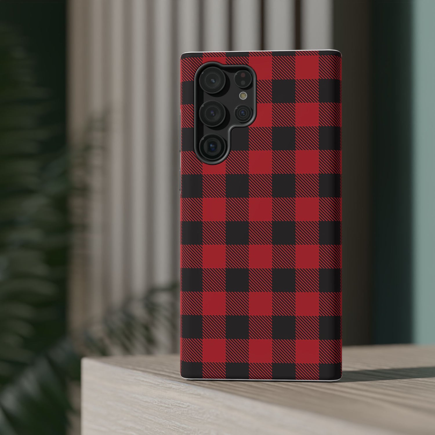 A red and black checkered phone case with a camera module on the back, placed on a wooden surface with a blurred plant in the background.