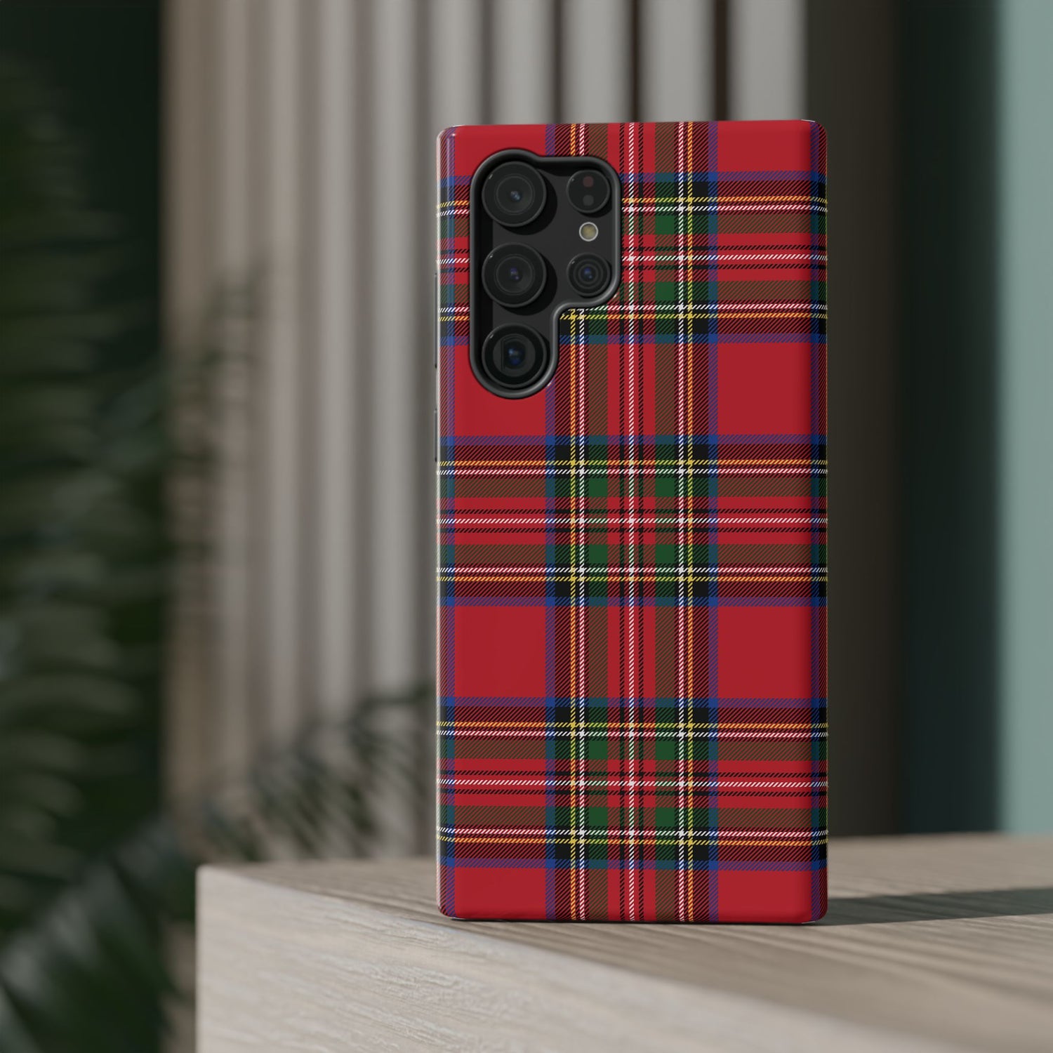 A red and green plaid patterned phone case with a camera module on the back, placed on a wooden surface with a blurred background.