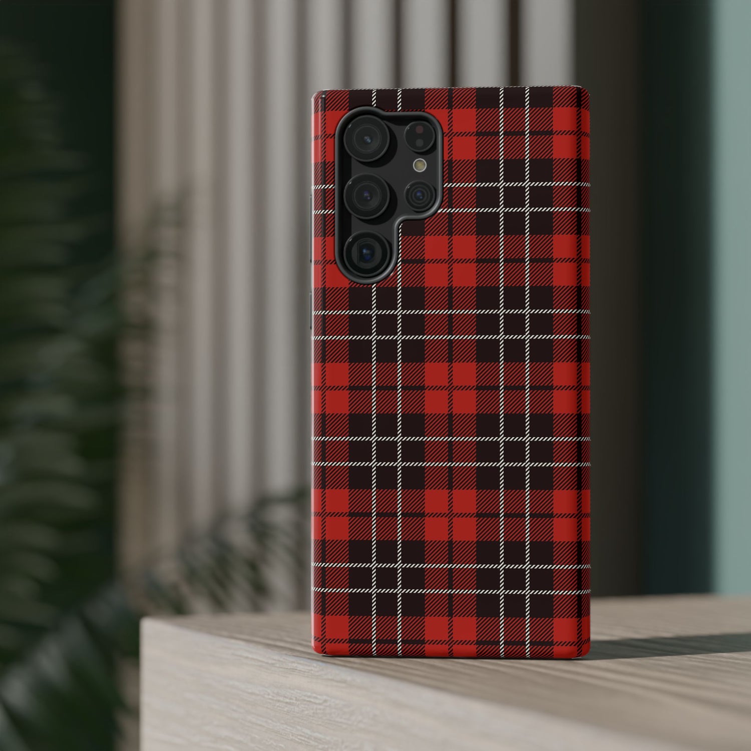A red and black checkered phone case with a camera module on the back, placed on a wooden surface with a blurred plant in the background.
