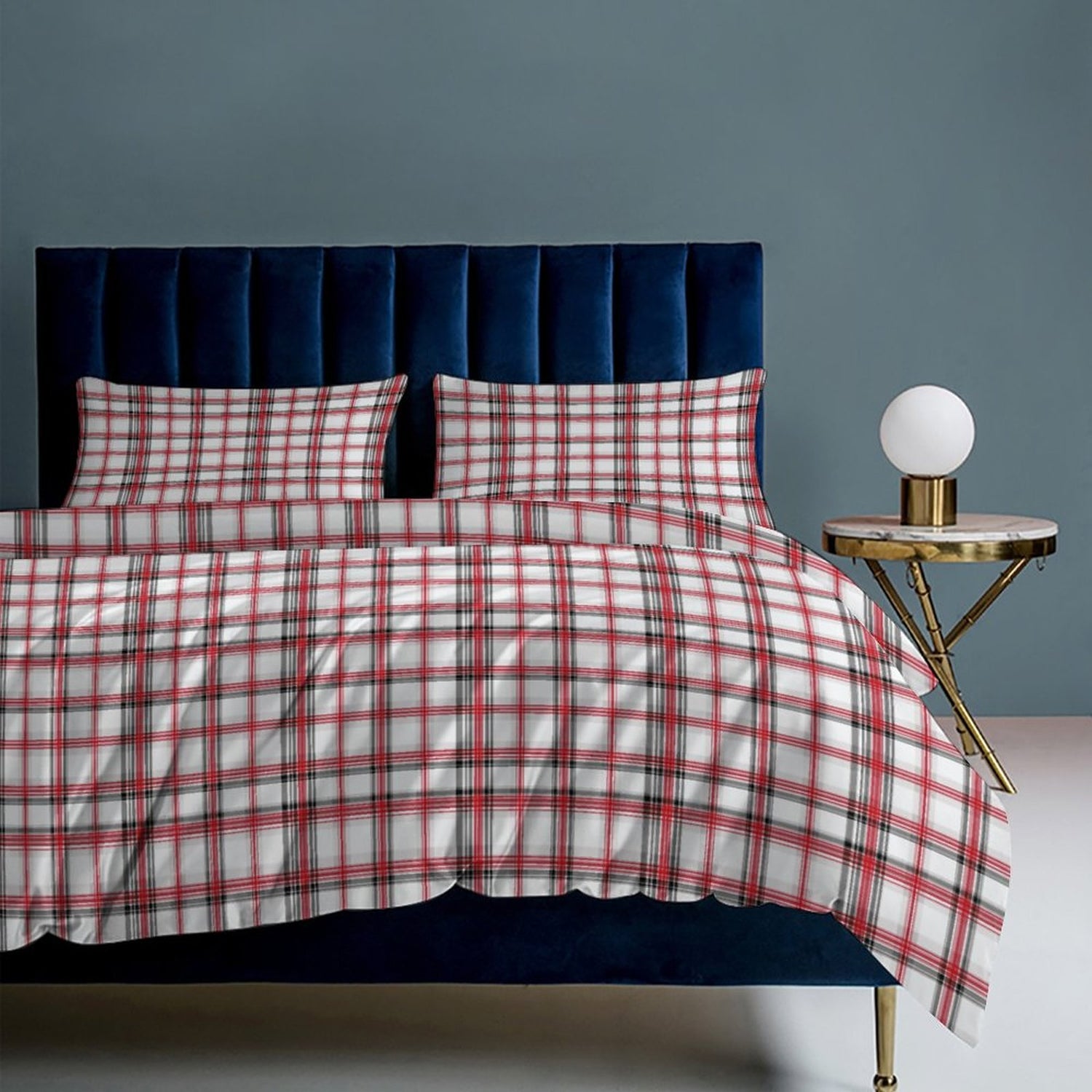 Plaid bedspread on a bed with a dark blue headboard and a small round table with a lamp in the corner.