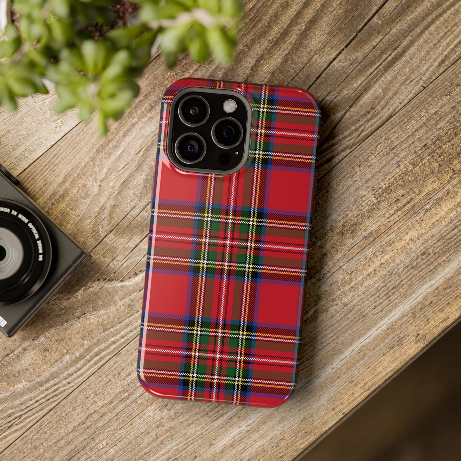 A red and green plaid patterned phone case with a camera module on the back, placed on a wooden surface next to a small camera.